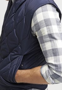 Chaleco acolchado azul marino con cierre frontal, que presenta una textura suave y brillante. Lleva puesto una camisa de franela blanca y azul a cuadros con los mangas arremangadas.