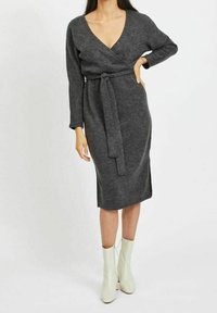 Šedé zavinovací šaty s dlouhými rukávy, výstřihem do V, uvázáním v pase a délkou ke kolenům. Nosené s bílými kotníkovými botami, které mají hladkou strukturu.