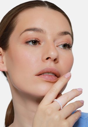 Femme avec un maquillage léger et des yeux marron touchant doucement ses lèvres, portant une bague en perles et un haut bleu.