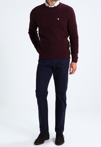 Viininpunainen ribbineulottu neulepaita yhdistettynä tummansinisiin chinohousuihin. Neuleessa on pyöreä pääntie ja hillitty kirjailtu logo rinnassa.