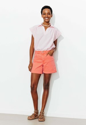 Femme debout et souriante portant un chemisier rayé clair, un short corail, un collier de coquillages et des sandales imprimé léopard, posant devant un mur blanc.