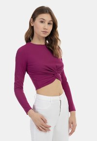 Tæt siddende, ribbet langærmet crop top i lilla med en knyttet frontdetalje. Kombineret med højtaljede hvide jeans, der viser et slankt design.