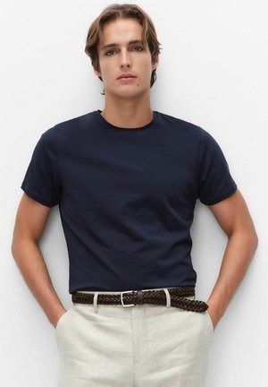 Junger Mann trägt ein dunkelblaues T-Shirt, das in beige Hosen mit braunem geflochtenem Gürtel gesteckt ist, steht mit den Händen in den Taschen vor weißem Hintergrund.