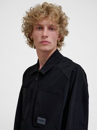 Jong persoon met krullend blond haar, die gouden creolen draagt en een zwarte jas met rits en voorzakken tegen een effen achtergrond.