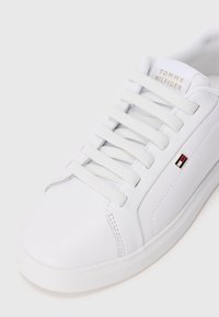 Baltos odinės sportbačiai su baltais raišteliais, "Tommy Hilfiger" logotipas ant liežuvio ir mažas raudonas, baltas ir mėlynas vėliavėlės ženklas šone.