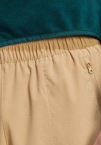 Beige elastische Hosen mit seitlichem Reißverschluss, kombiniert mit einem strukturierten dunkelgrünen Oberteil aus glattem, weichem Stoff.