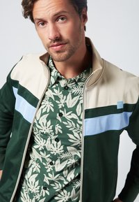 Gr ön zip-up jacka med beige, mörkgröna och ljusblå ränder. Underneath, en grön blommönstrad skjorta. Slät tygtextur.