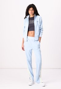 Lys blå treningsdrakt med glidelåsjakke og matchende bukser. Den har hvite side-striper og en svart crop-top under. Glatt tekstur.