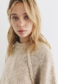 Béžový pletený sveter s vysokým ribovaným golierom a voľným strihom. Textúra vyzerá mäkko, s kombináciou jemných a hrubých niťí.