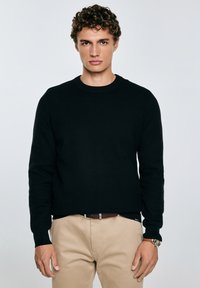 Un giovane con capelli ricci indossa un maglione nero lavorato e pantaloni beige, in piedi davanti a uno sfondo chiaro e semplice.