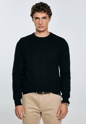 En ung mand med krøllet hår har en sort struktureret sweater og beige bukser på, mens han står foran en ensfarvet lys baggrund.