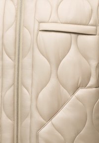 Tissu matelassé beige avec des surpiqûres en forme de sablier, une fermeture éclair métallique verticale sur la gauche et deux ouvertures de poche horizontales ainsi qu'une en biais.