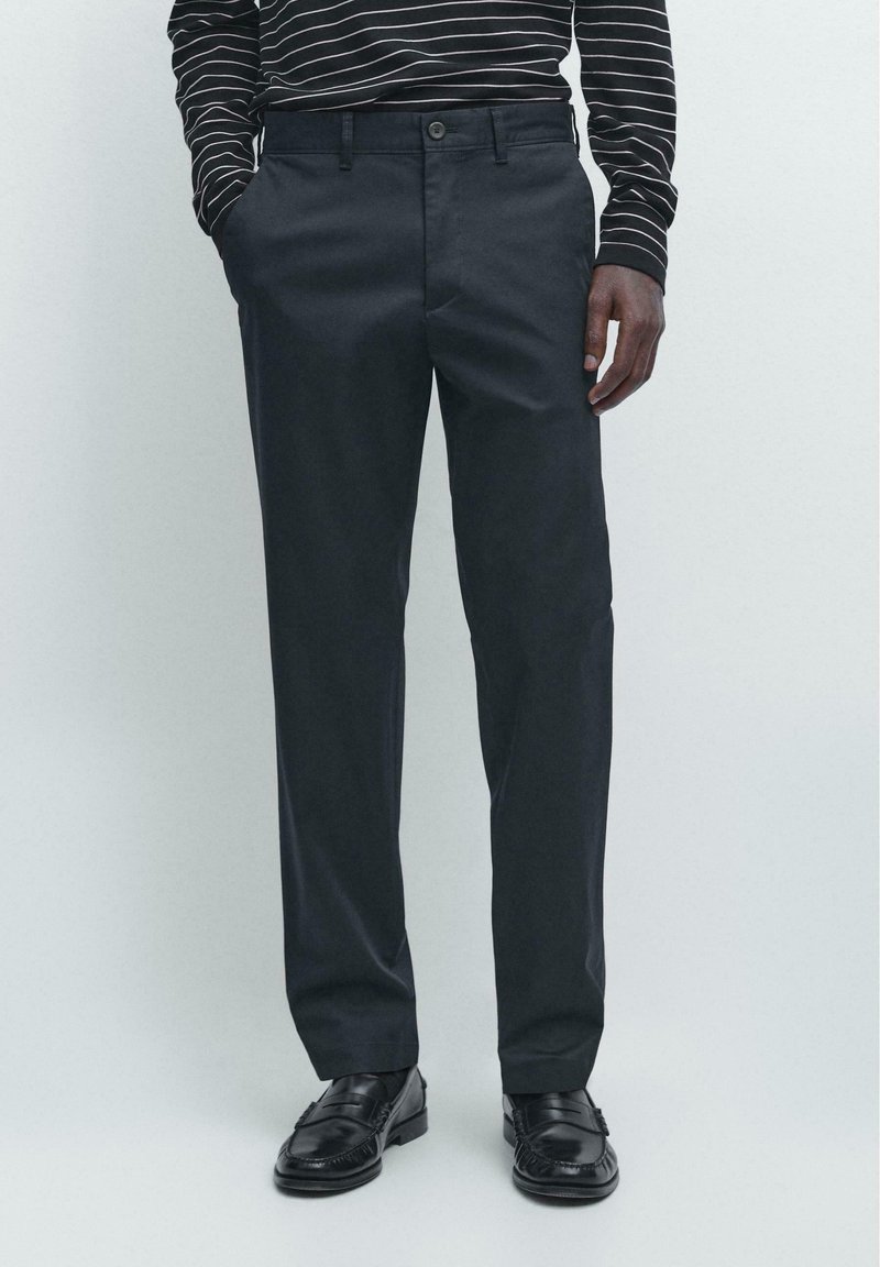 Uomo con pantaloni neri a gamba dritta, mocassini neri e una camicia nera a maniche lunghe con righe orizzontali bianche.