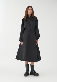 Camisa negra de manga larga con botones combinada con una falda midi negra texturizada que presenta un patrón de diamantes y botas negras hasta el tobillo.