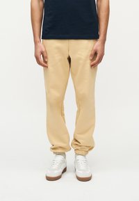 Άνδρας που φοράει μπεζ παντελόνια αθλητικού τύπου, λευκά αθλητικά παπούτσια και σκούρο μπλε πουκάμισο, που στέκεται μπροστά σε λευκό απλό φόντο.