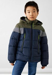 Veste rembourrée avec capuche en marine, vert et gris. Elle dispose de deux poches avant zippées et de poignets élastiques pour un ajustement confortable.