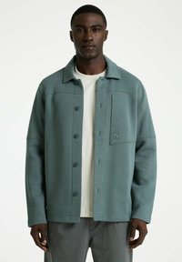 Getailleerde groene jas van gestructureerde stof, met een knoopsluiting aan de voorkant, kraag en borstzak met logo-detail. Draagt over een licht shirt.