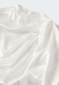 Blouse blanche en satin avec un col montant et une fermeture éclair au dos, présentant un drapé léger du tissu et des manches légèrement bouffantes.