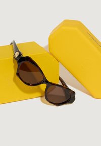 Óculos de sol em padrão tartaruga com lentes castanhas, apresentando uma armação brilhante e casos texturizados amarelos distintos, dispostos em ângulo para exibição.