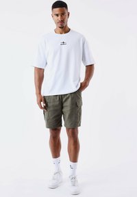Camiseta blanca de manga corta con logotipo negro, pantalones cortos de cargo oliva, zapatillas blancas y calcetines blancos con estampado. Conjunto casual con un ajuste cómodo.