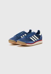 Navy blue sneakers with white stripes, suede material, rubber sole, red accent on midsole, and round laces. Features textured panels.