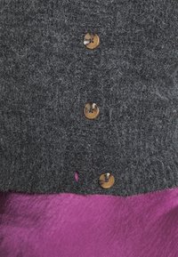 Tmavě šedý svetr s texturou, hnědými knoflíky a malým otvorem. Položený na živé fialové látce. Důraz na materiál a detaily knoflíků.