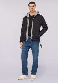 Chaqueta de plumas negra con capucha gris, que presenta un diseño acolchado, cierre con cremallera y bolsillos laterales. Combinada con jeans de mezclilla azules y zapatillas blancas.