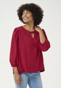 Red textured blouse with a rounded neckline, keyhole detail, and drawstring tie. Features puff sleeves and a loose-fitting design.