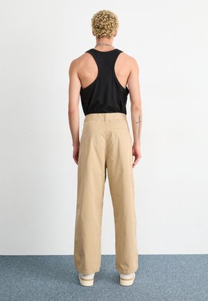 Person with curly blond hair wearing a black tank top, beige wide-leg pants, white shoes, and a sword tattoo on their left forearm, standing.