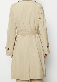 Trench coat beige con vita allacciata, maniche lunghe e bottoni sui polsini; tessuto liscio con una leggera lucentezza e un design classico.