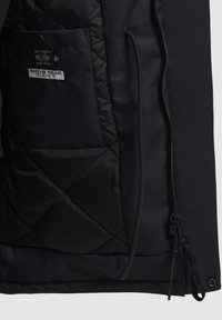 Chaqueta negra acolchada con cremallera frontal, un bolsillo parche y una etiqueta de tela con texto. Material exterior liso y duradero.