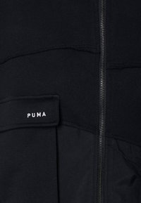 Čierna zipová mikina vyrobená z mäkkej textílie, so štítkom PUMA, kontrastným šitím a hladkým zipsom na prednej strane.