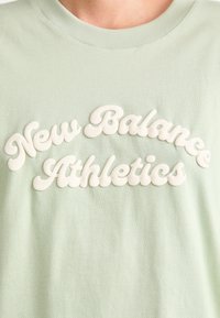 T-shirt en coton vert clair avec un texte en relief de couleur crème qui dit "New Balance Athletics" dans une police ondulée.