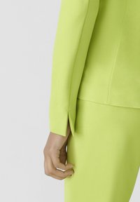 Veste sur mesure vert lime au design élégant, dotée d'une fente au niveau du poignet et d'une texture lisse. Main positionnée près de l'ourlet.