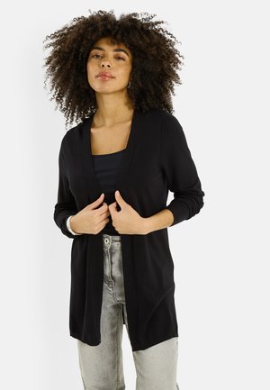 Mujer con cabello rizado que lleva un cárdigan negro abierto sobre una blusa negra y jeans gris claro, de pie contra un fondo blanco.