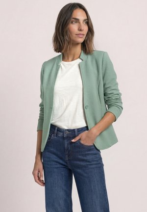 Kvinde med skulderlangt brunt hår iført en lysegrøn blazer, hvid t-shirt og blå jeans med den ene hånd i lommen, ser sidelæns.