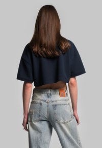 Morska skurzona bluza z szerokim dekoltem, zestawiona z jasnoniebieskimi, wysokimi dżinsami. Skórzana metka marki na pasku, z wyblakłą teksturą dżinsu.