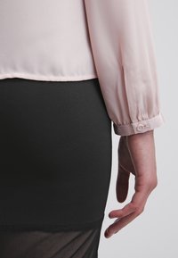 Blusa trasparente rosa chiaro con dettagli sui polsini, infilata in una gonna nera aderente con sovrapposizione in rete all'orlo. Contrasti di texture sottili.