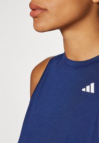 Marineblå ærmeløs atletisk top med høj krave, der har et hvidt geometrisk logo øverst til venstre. Blød stoftekstur.