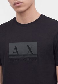 Άνδρας που φοράει μαύρο t-shirt Armani Exchange με υφή λογότυπου AX και όνομα μάρκας μπροστά, εμφανιζόμενος από τους ώμους μέχρι το μέσο του στήθους.