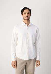 Witte blouse met lange mouwen, knoopsluiting aan de voorkant, kraag en een gladde textuur. Draagt over beige shorts met een gebogen zoom.