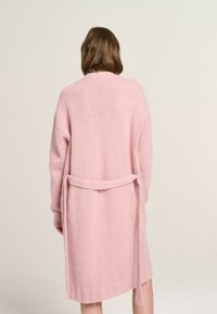 Langer pinkfarbener Cardigan aus weichem, strukturiertem Stoff. Verfügt über überschnittene Schultern, einen Bindegürtel in der Taille und gerippte Ärmel- und Saumdetails.