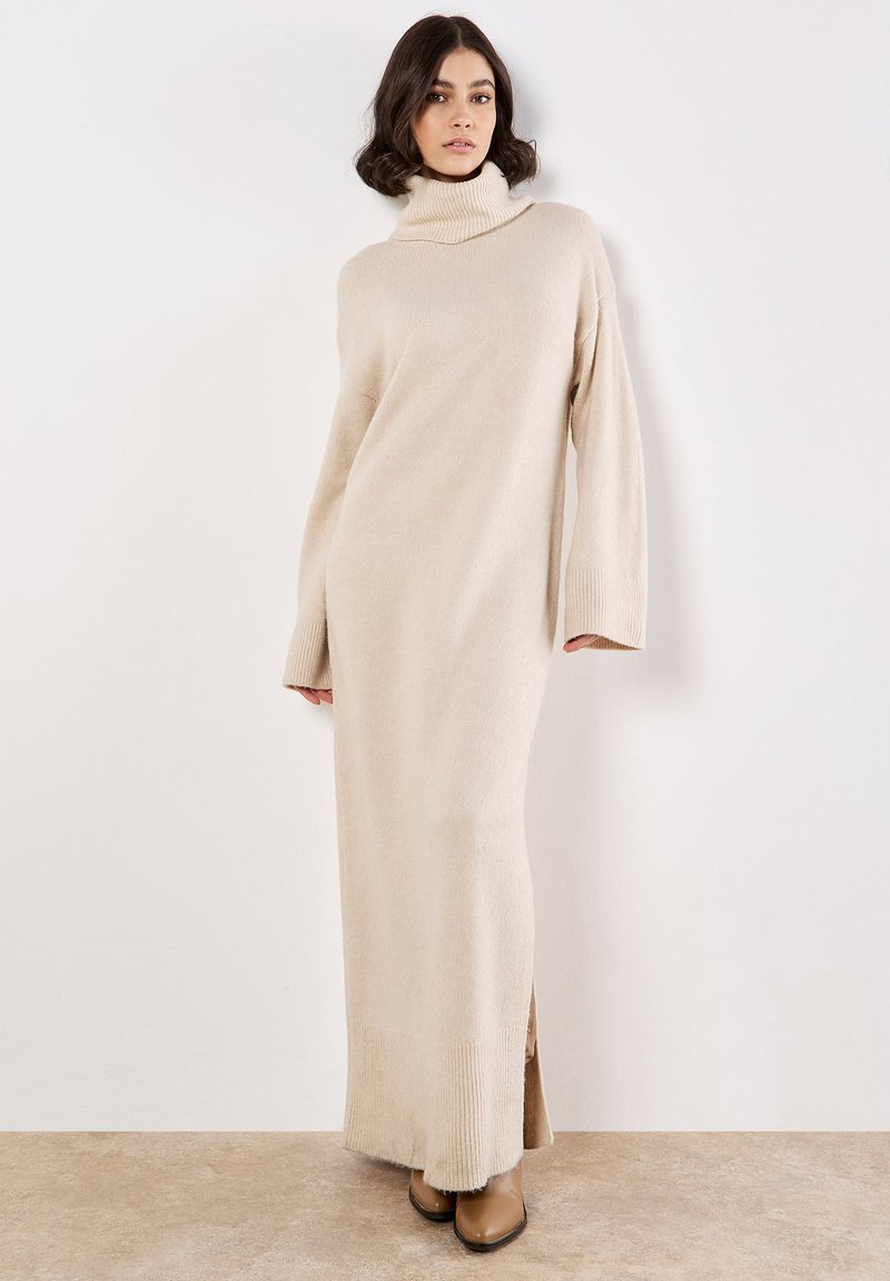 Robe tricotée surdimensionnée couleur crème avec un col montant, des manches longues et des fentes sur les côtés. Texture douce et accents côtelés aux poignets et à l'ourlet.