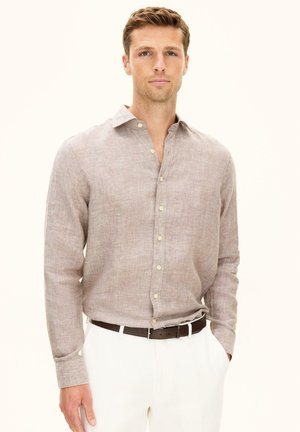 Mann trägt ein hellbraunes, langärmeliges Hemd mit Knöpfen, das in weiße Hosen mit einem dunklen Gürtel gesteckt ist, steht mit einer Hand in der Tasche.