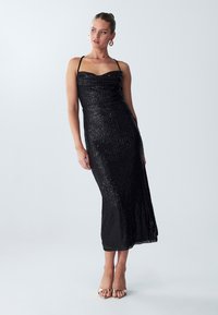 Abito lungo nero con pailettes, scollatura a cuore, spalline sottili e silhouette aderente. Tessuto liscio, spacco all'altezza del ginocchio, indossato con sandali dorati.