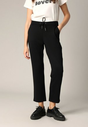 Pantalon noir ajusté avec taille à cordon, poches latérales et texture lisse, associé à des chaussures noires à lacets et un t-shirt graphique blanc.