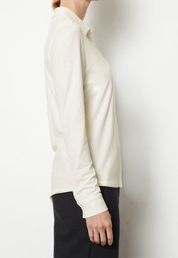 Femme portant une chemise à col longue à manches crémeux et un pantalon noir, se tenant de profil contre un fond blanc uni.