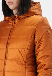 Gevoerde jas in een levendige oranje kleur, met een ritssluiting en een capuchon. Kenmerkt zich door horizontale stiksels en een rond logo op de mouw.