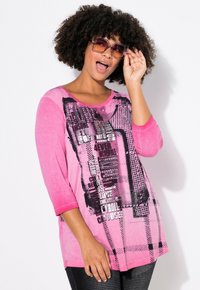 Haut à manches longues rose clair avec texte graphique noir et motifs, arborant une coupe décontractée et un col rond, associé à des leggings noirs texturés.