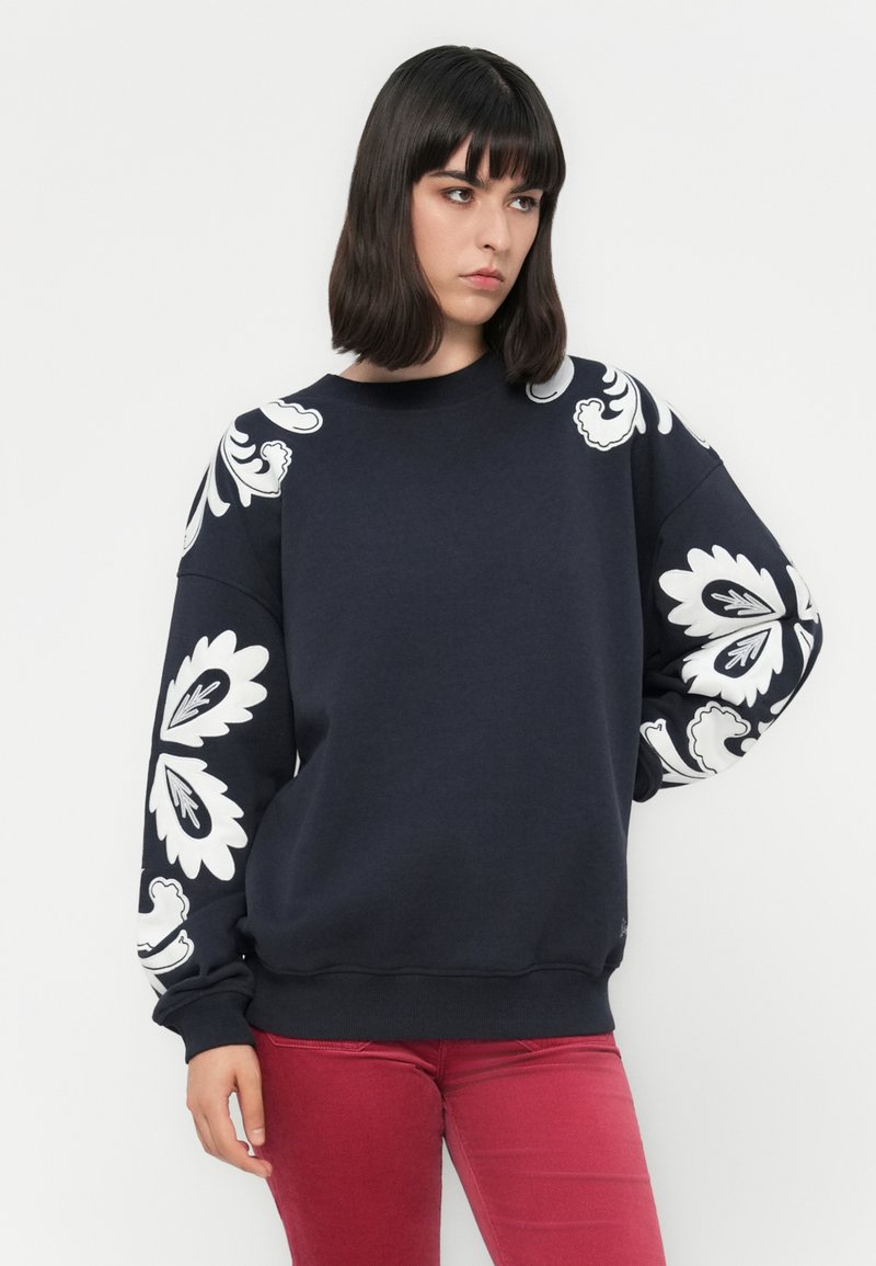 Marineblauwe sweatshirt met witte bloemmotieven op de mouwen. Heeft een ronde halslijn en een relaxte pasvorm, gemaakt van zachte katoenen stof.